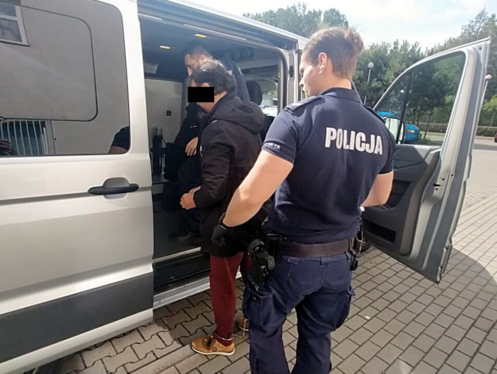 Funkcjonariusz policji eskortuje osobę do wnętrza vanu w otoczeniu parkingu.