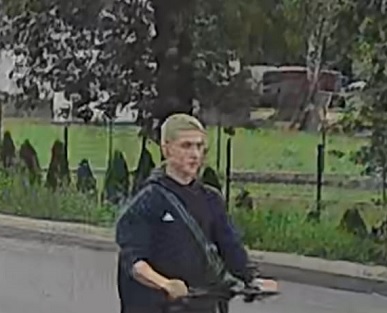Młody mężczyzna na hulajnodze porusza się po ulicy, w tle widać zieloną przestrzeń i ogrodzenie.