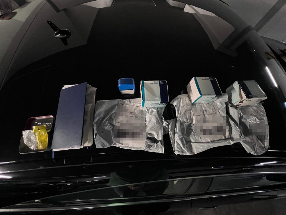 Na czarnej powierzchni samochodu znajdują się różne opakowania, dokumenty oraz małe przedmioty, w tym plastikowy pojemnik.