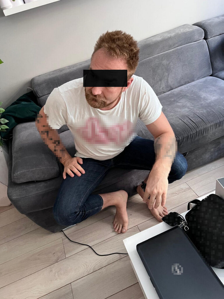 Mężczyzna w białym t-shircie siedzący na szarej kanapie, z nogami skrzyżowanymi, obok laptopa i torby.