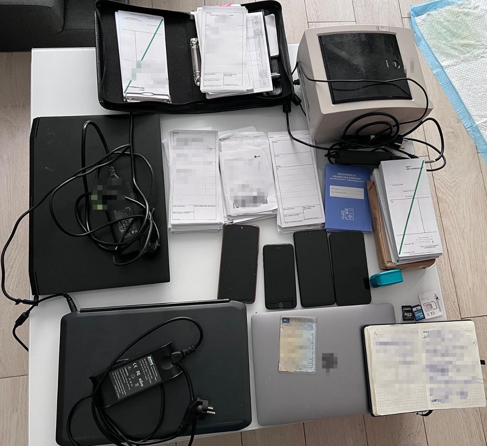 Na biurku rozłożone są dokumenty, dwa laptopy, kilka telefonów, drukarka oraz różne akcesoria elektroniczne.