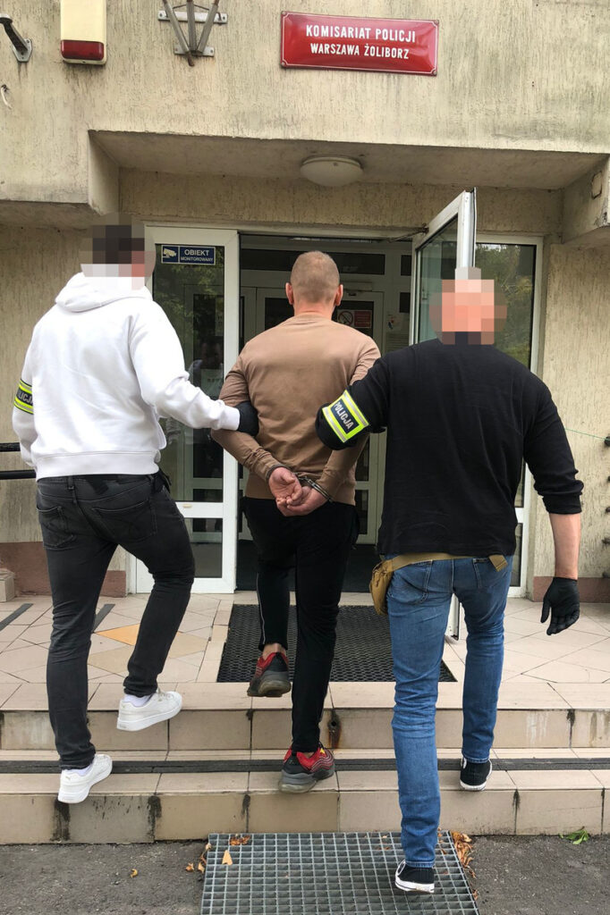 Mężczyzna w kajdankach prowadzony przez dwóch policjantów do wejścia do komisariatu.