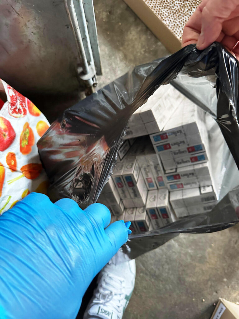 Osoba w niebieskiej rękawiczce trzyma czarną torbę, z której widać paczki papierosów na tle posadzki.