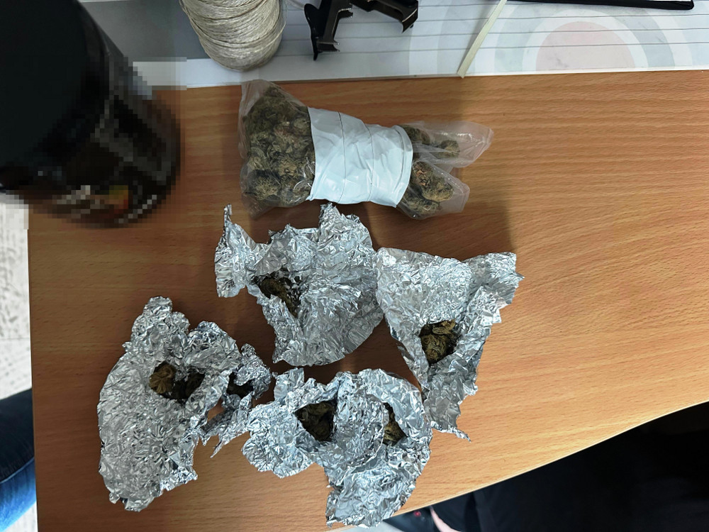 Na stole leżą cztery zawiniątka w folii aluminiowej oraz jeden pakunek w białym papierze, zawierające nieokreślony materiał roślinny.
