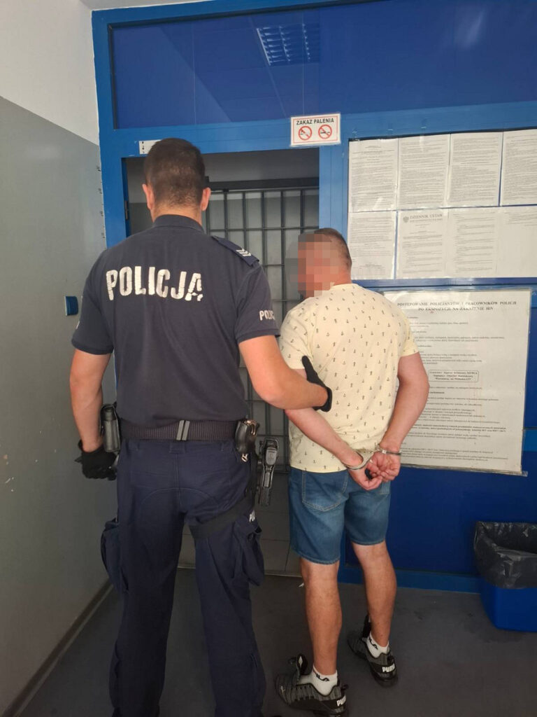 Policjant w mundurze prowadzi zatrzymanego mężczyznę z rękami związanymi z tyłu w korytarzu komisariatu.