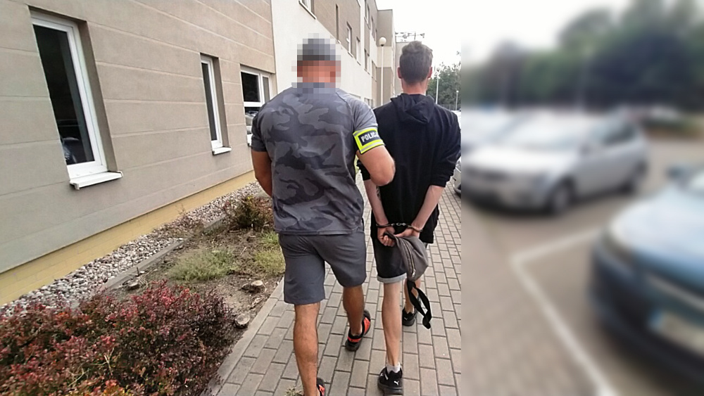 Mężczyzna w mundurze policyjnym eskortuje aresztowanego mężczyznę wzdłuż budynku.