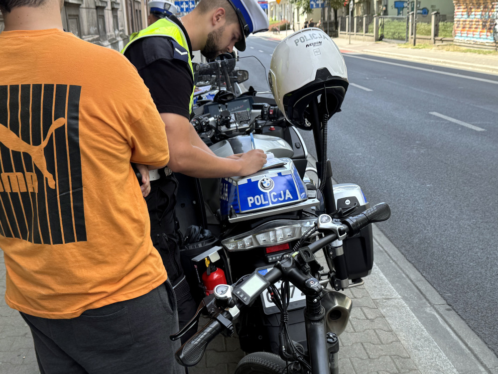 Policjant w mundurze zapisuje notatki przy radiowozie, obok stoi mężczyzna w pomarańczowej koszulce.