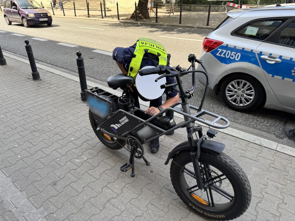Policjant w kamizelce odblaskowej sprawdza elektryczny rower na chodniku obok policyjnego samochodu.