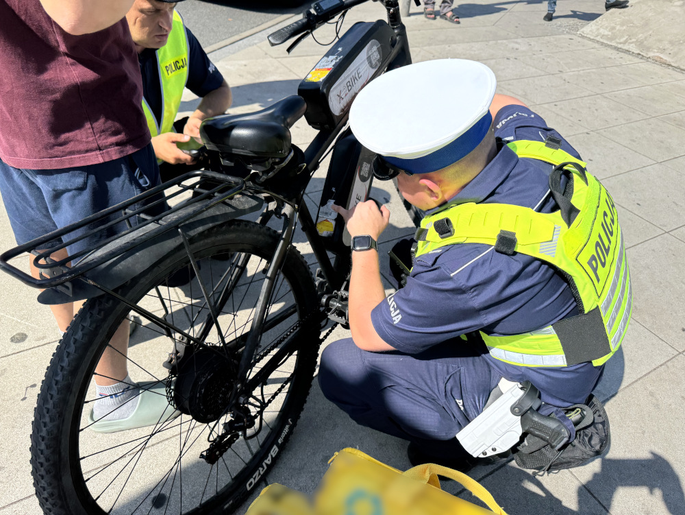 Policjant w odblaskowej kamizelce naprawia elektryczny rower na chodniku, obok stoi młody mężczyzna.