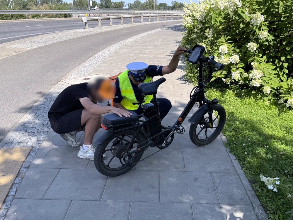 Mężczyzna w czarnym t-shircie i krótkich spodniach klęczy obok elektrycznego roweru, a policjant w kamizelce odblaskowej sprawdza pojazd. W tle widać kwitnące krzewy oraz drogę.