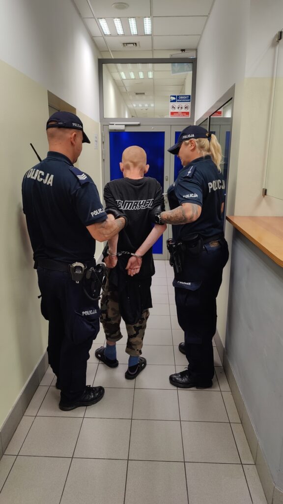 Mężczyzna w kajdankach prowadzony przez dwóch policjantów w korytarzu budynku.