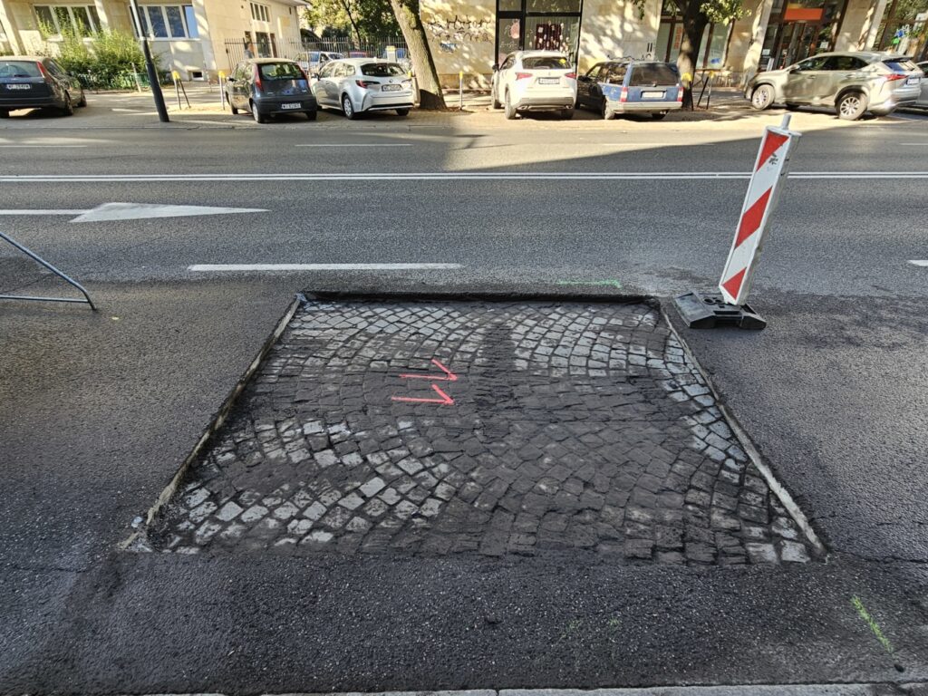 Remontowany fragment chodnika z kostki brukowej, oznaczony różowymi liniami, obok ruchliwej ulicy.