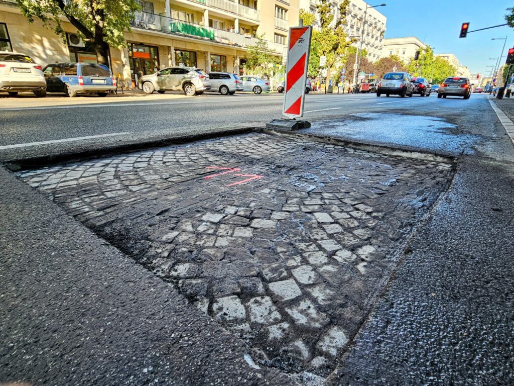 Kostka brukowa w wykopie drogowym, oznaczona różową farbą, obok ruchliwej ulicy z samochodami i sygnalizacją świetlną.
