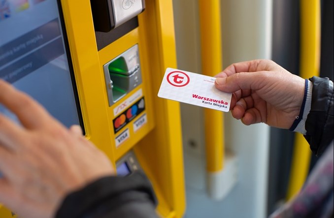 Osoba trzyma kartę miejską przy automacie biletowym w tramwaju, przygotowując się do zakupu biletu.