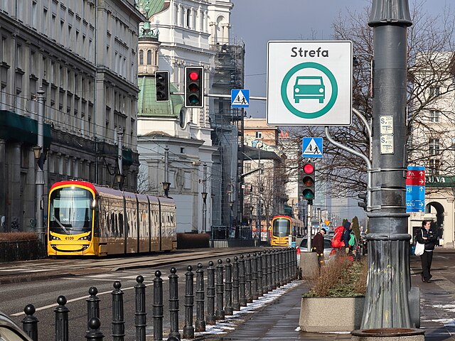 Tramwaje w żółtej kolorystyce zatrzymują się na skrzyżowaniu, obok znaku "Strefa", w miejskim otoczeniu.