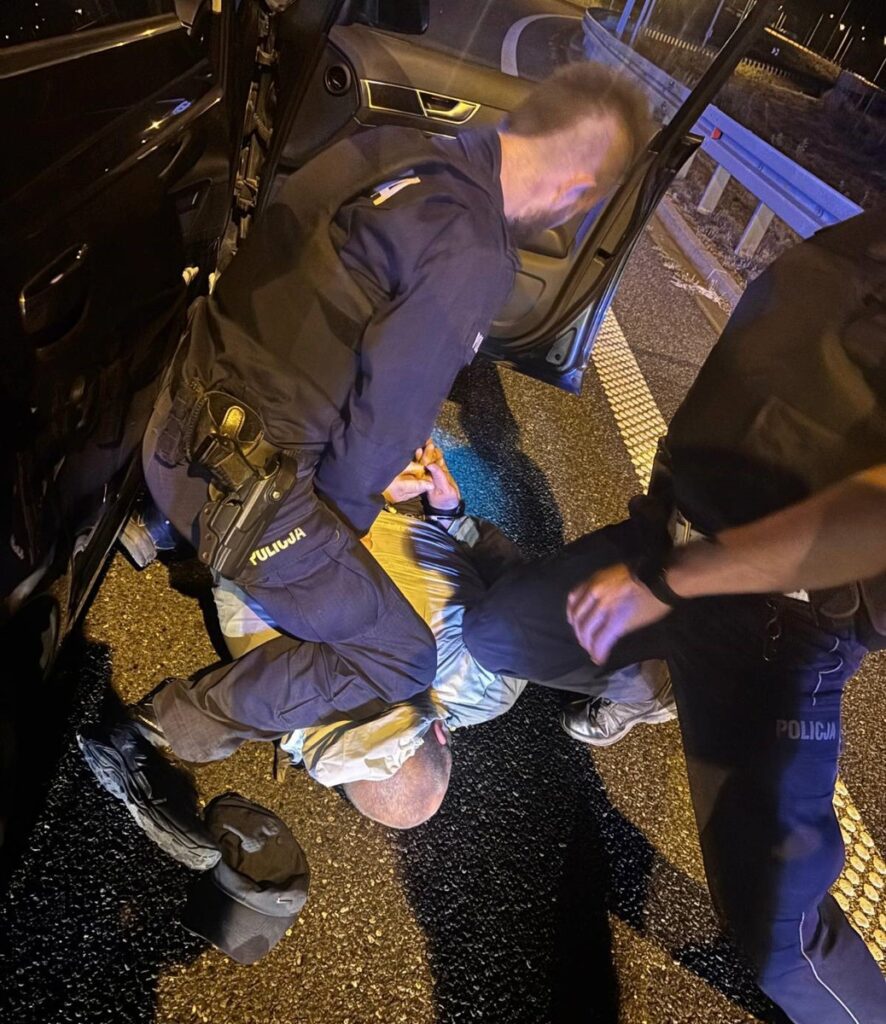 Dwaj policjanci zatrzymują mężczyznę leżącego na jezdni, jeden z nich trzyma go za ręce, a drugi stoi obok.