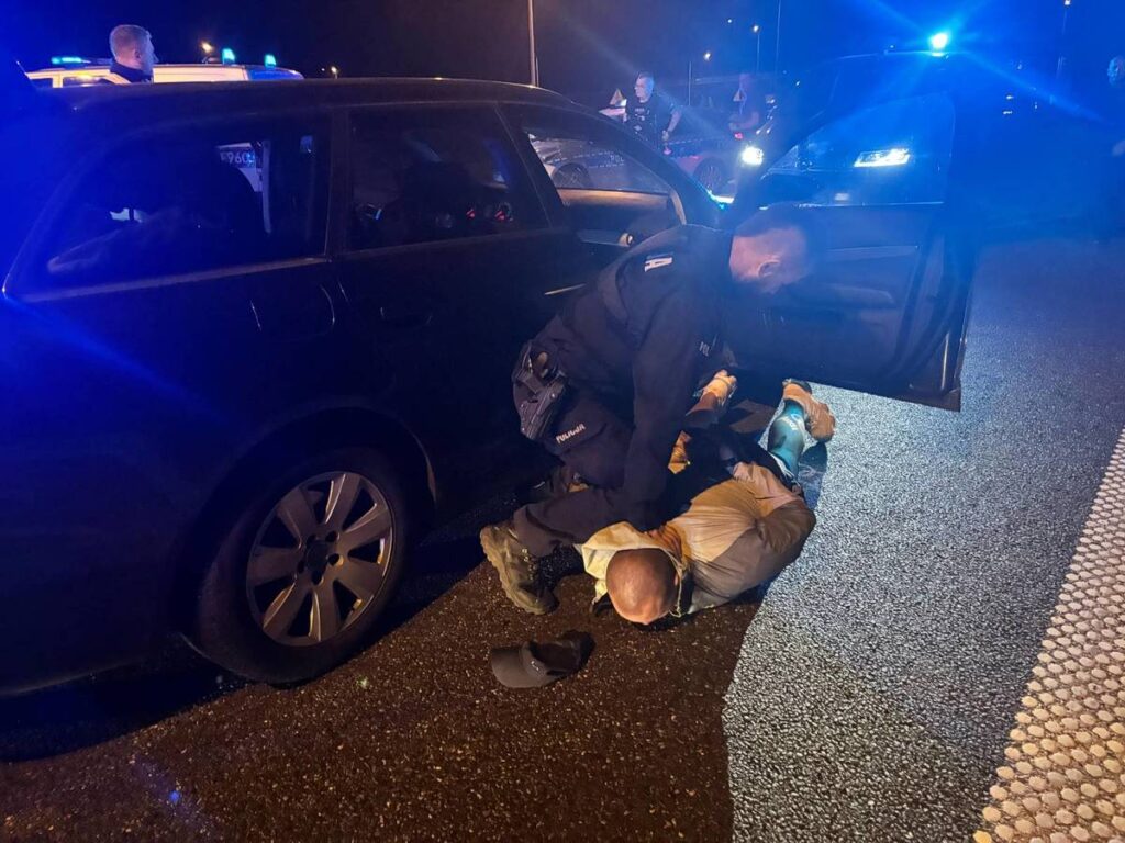 Policjant obezwładnia mężczyznę leżącego na drodze obok zaparkowanego samochodu w nocy, w otoczeniu radiowozów.