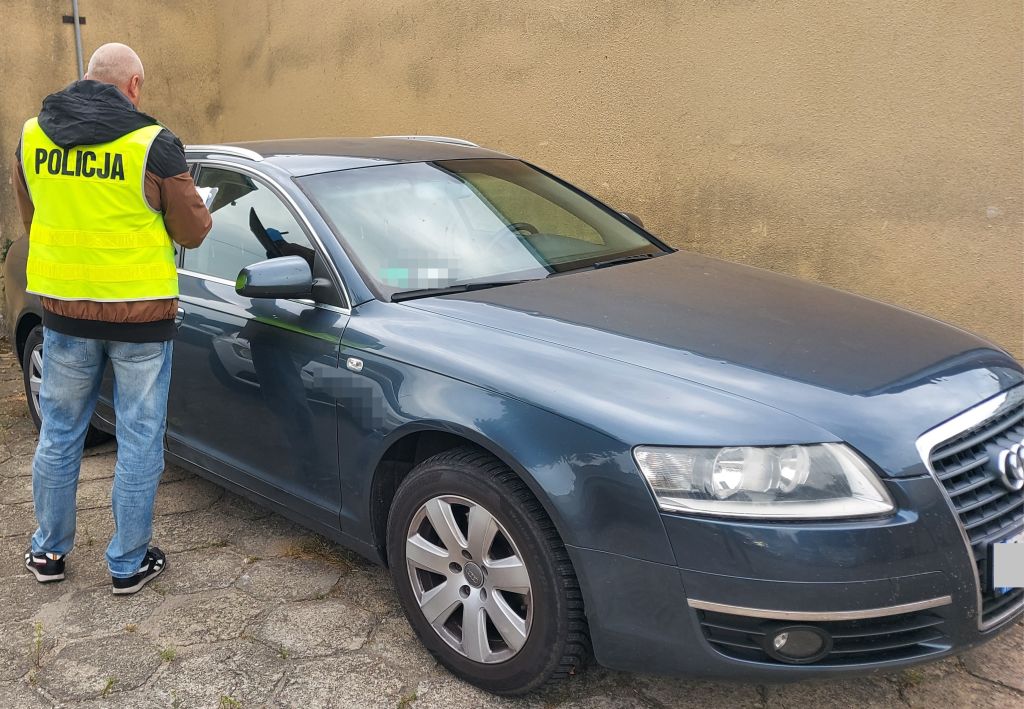Policjant w kamizelce odblaskowej sprawdza wnętrze niebieskiego samochodu osobowego zaparkowanego przy ścianie.