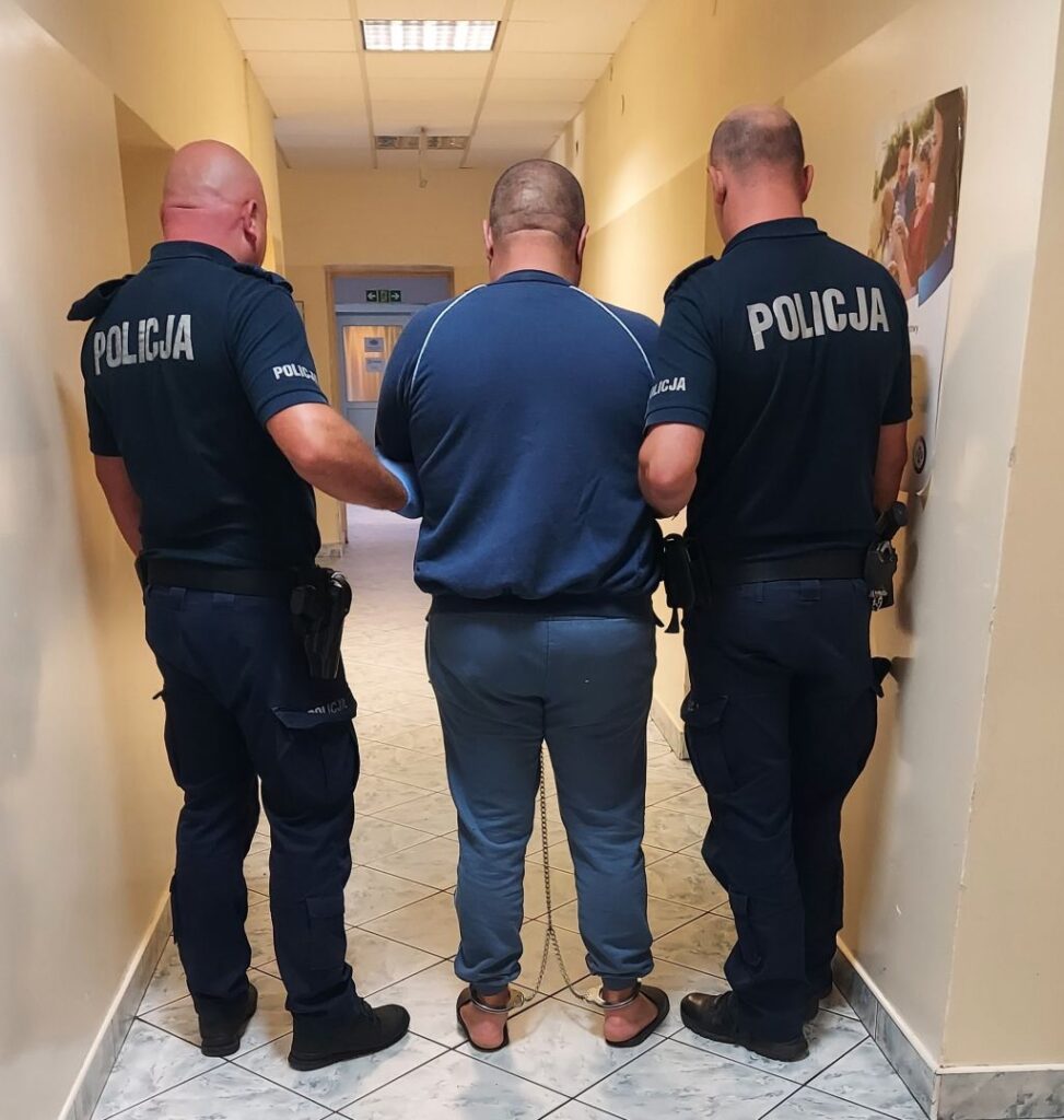 Mężczyzna w kajdankach prowadzony przez dwóch policjantów w korytarzu budynku.