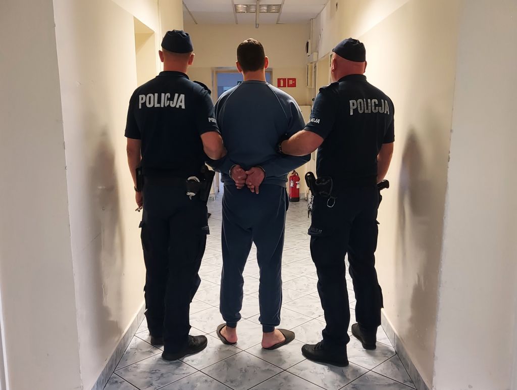 Mężczyzna w dresie z rękami związanymi z tyłu prowadzony przez dwóch policjantów w korytarzu budynku.