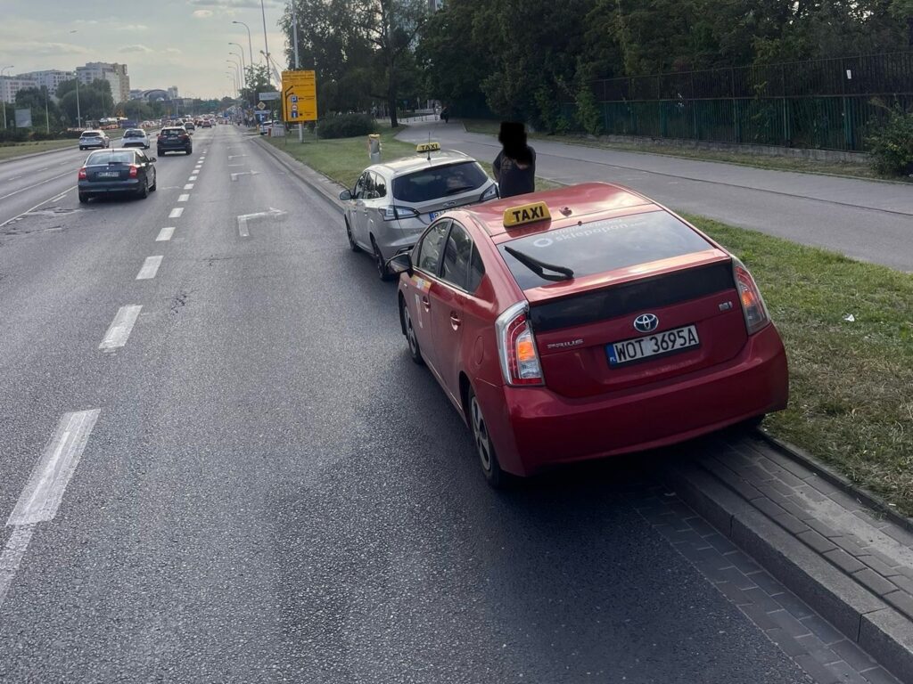 Czerwony taksówka zaparkowana na poboczu drogi, obok przejeżdżające samochody.