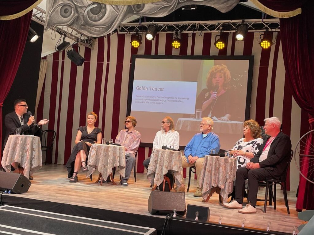 Mężczyzna w garniturze prowadzi dyskusję z grupą osób siedzących przy stołach na scenie w sali konferencyjnej.