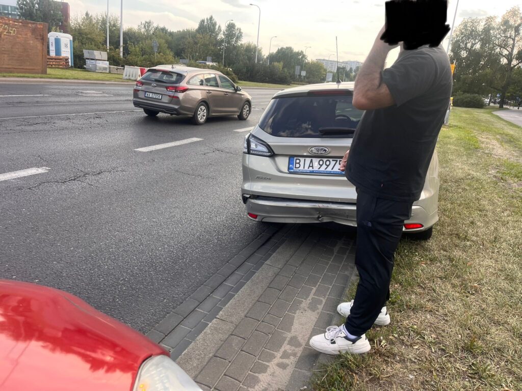 Mężczyzna w czarnej koszulce rozmawia przez telefon obok uszkodzonego samochodu na poboczu drogi.