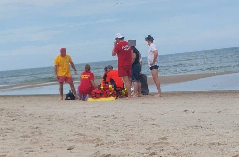 Grupa ratowników i osób postronnych udziela pomocy poszkodowanemu na plaży, obok wody.