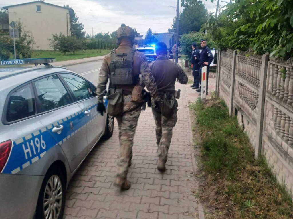 Dwóch mężczyzn w mundurach wojskowych idzie chodnikiem obok radiowozu policyjnego w miejskim otoczeniu.