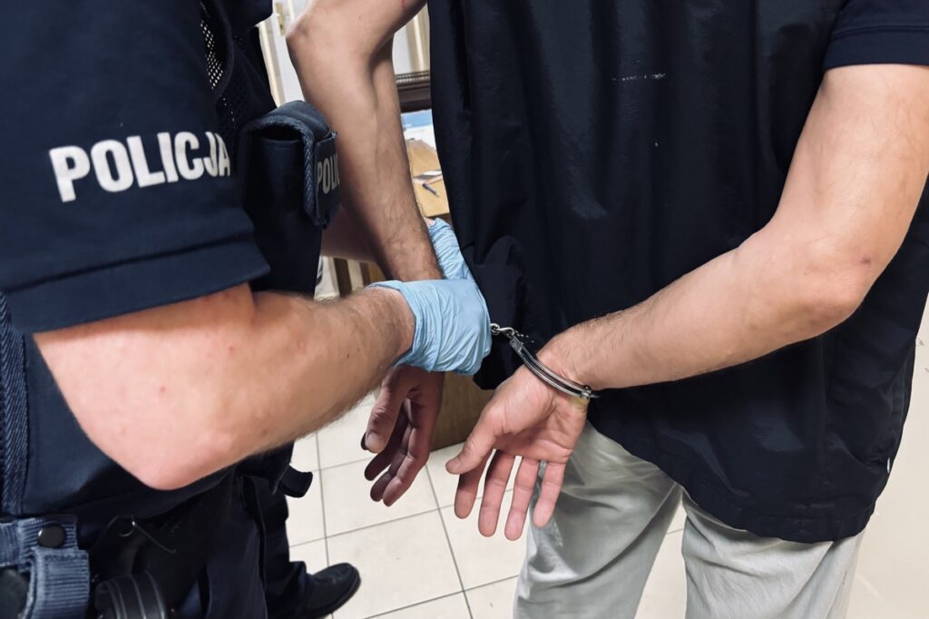 Policjant zakłada kajdanki mężczyźnie w czarnej koszuli w pomieszczeniu z jasną podłogą.