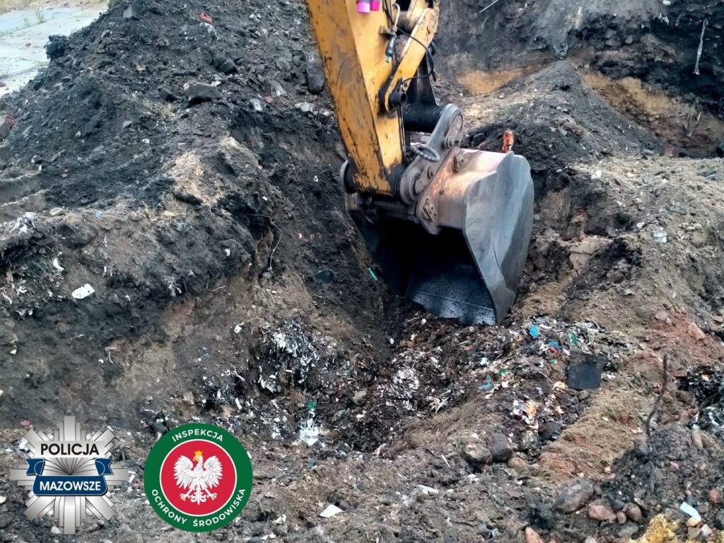 Koparka wydobywająca odpady z wykopu w zanieczyszczonym terenie. W tle widoczne hałdy ziemi i śmieci.