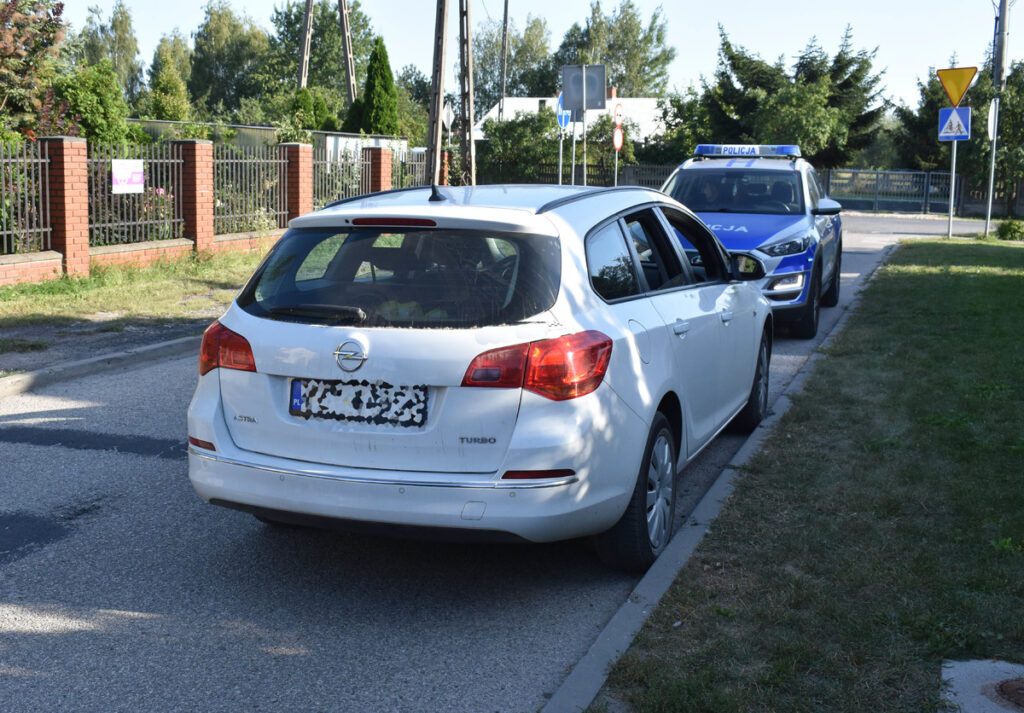 Biały samochód osobowy zaparkowany na poboczu drogi obok nieoznakowanego radiowozu policyjnego.