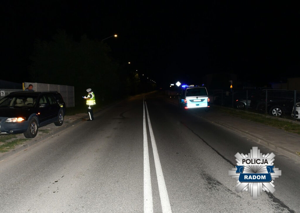 Policjant w odblaskowej kamizelce kontroluje dokumenty kierowcy przy zatrzymanym samochodzie na oświetlonej ulicy.