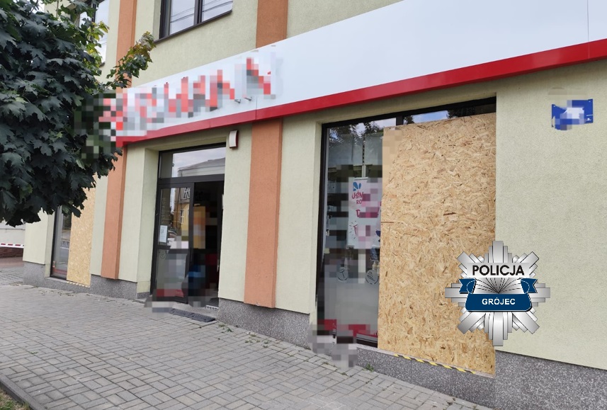 Zamknięty lokal usługowy z zablokowanym wejściem, zasłoniętym płytą OSB, w otoczeniu drzew i chodnika.