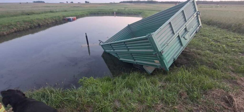 Zielona przyczepa leży na boku w wodzie, otoczona trawą i polami. W tle widoczne niskie wzgórza.
