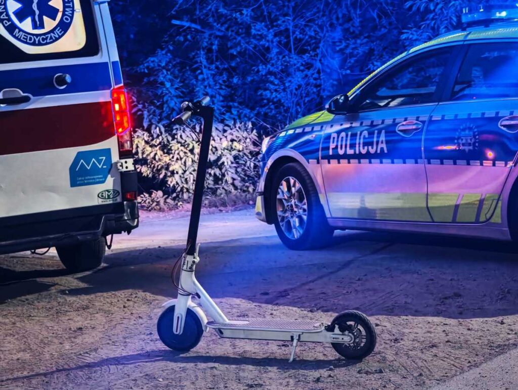 Skuter elektryczny stoi na drodze obok radiowozu policyjnego i karetki, otoczony niebieskim światłem.