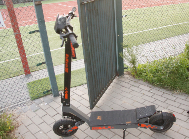 Czarna hulajnoga elektryczna stoi przy otwartych drzwiach na tle boiska sportowego.