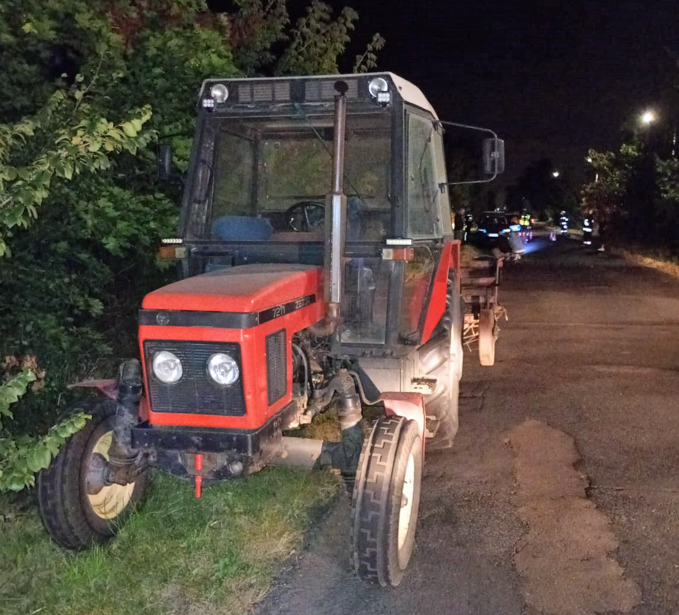 Traktor z czerwoną maską stoi na poboczu drogi nocą, otoczony przez zieleń i oświetlenie uliczne.
