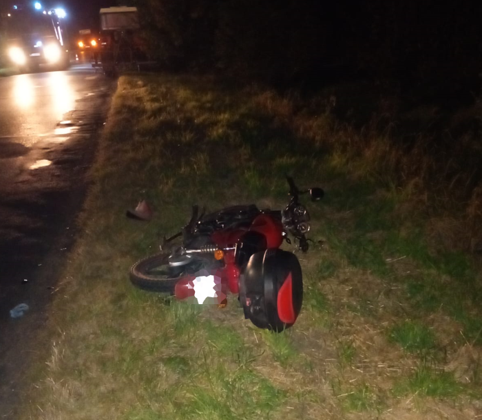 Czerwony motocykl leży na poboczu drogi, w tle widać pojazd z włączonymi światłami.
