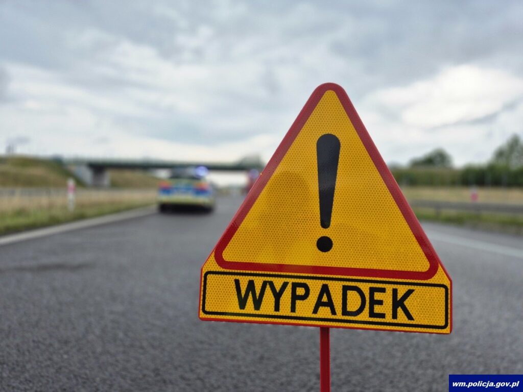 Na drodze stoi żółty znak ostrzegawczy z napisem "WYPADEK", w tle widoczny radiowóz policyjny.