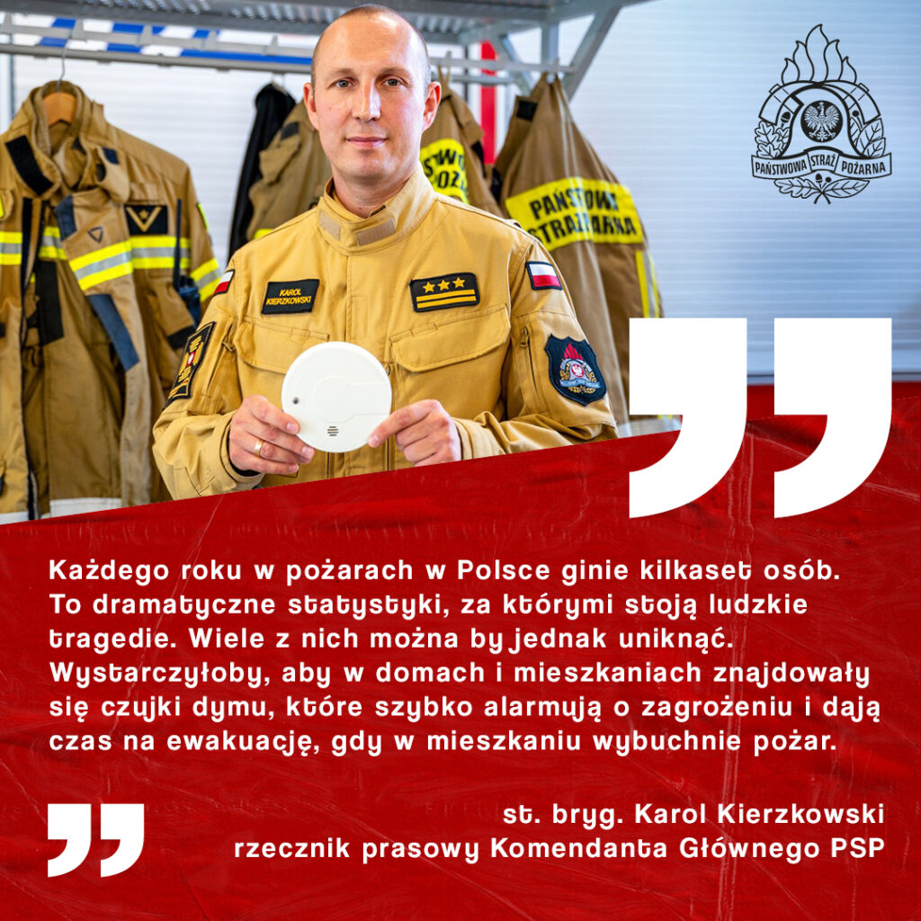 Mężczyzna w mundurze strażackim trzyma czujnik dymu, stojąc przed wieszakiem z odzieżą strażacką w tle.