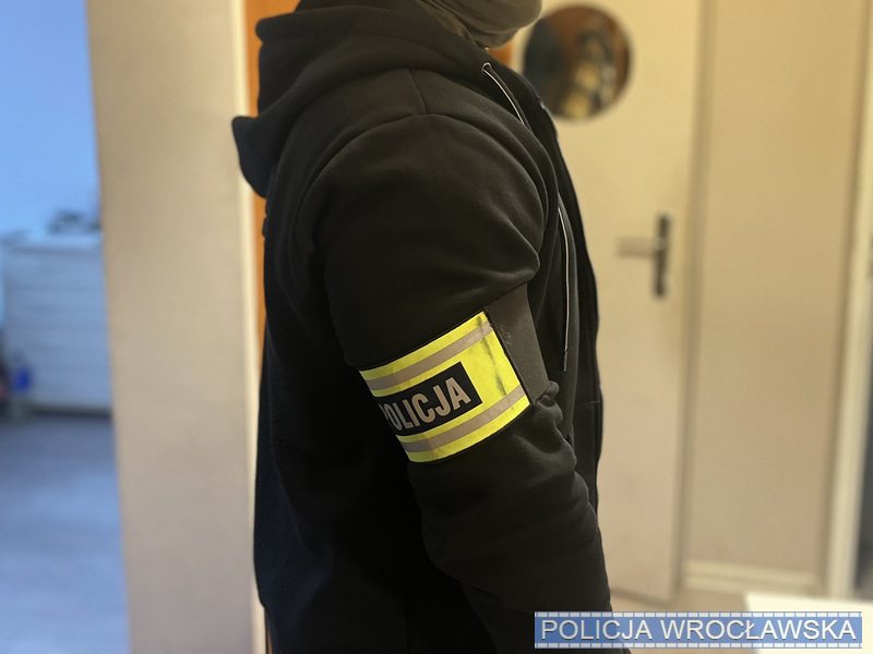Mężczyzna w czarnej bluzie z kapturem, z opaską "POLICJA" na ramieniu, stoi w pomieszczeniu z jasnymi ścianami.