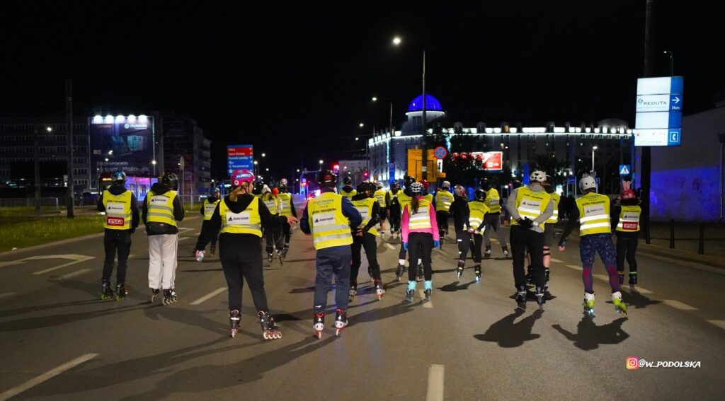 Grupa osób na rolkach porusza się po oświetlonej ulicy nocą, nosząc odblaskowe kamizelki.