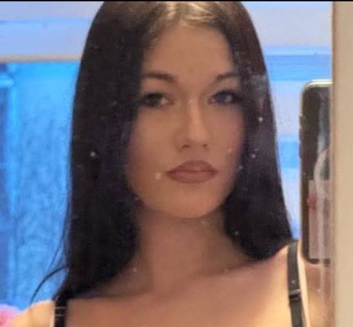 Kobieta z długimi, prostymi włosami robi sobie selfie w lustrze, na tle okna z niebieskim światłem.