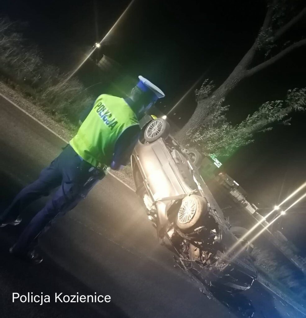 Policjant w kamizelce odblaskowej bada przewrócone auto na drodze w nocy, otoczone światłami.