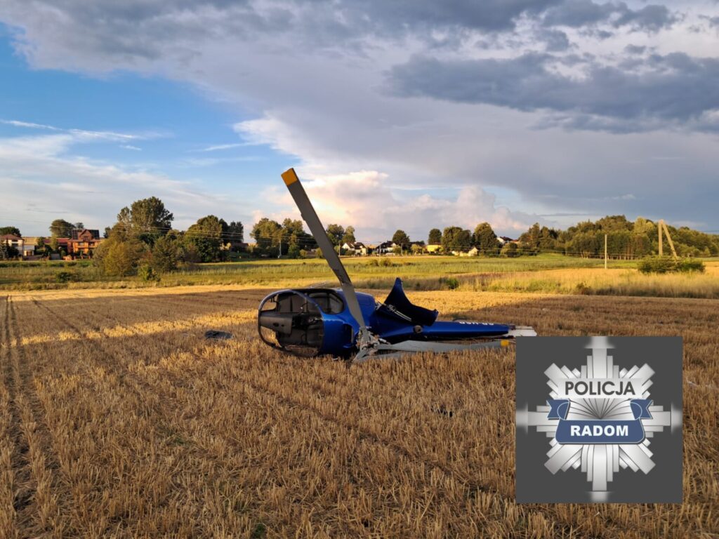 Helikopter leży na polu z zebranym zbożem, w tle widoczne domy oraz drzewa.