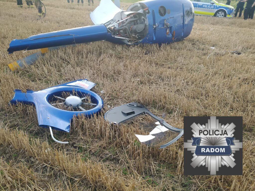 Rozbity śmigłowiec leży na polu, obok fragmenty wirnika i elementy konstrukcyjne, w tle widoczni funkcjonariusze.
