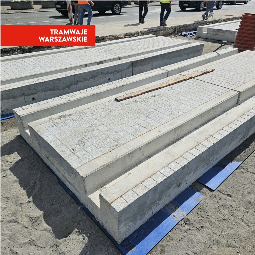 Betonowe elementy torowiska tramwajowego układane na placu budowy w mieście, otoczone przez pracowników w kamizelkach odblaskowych.