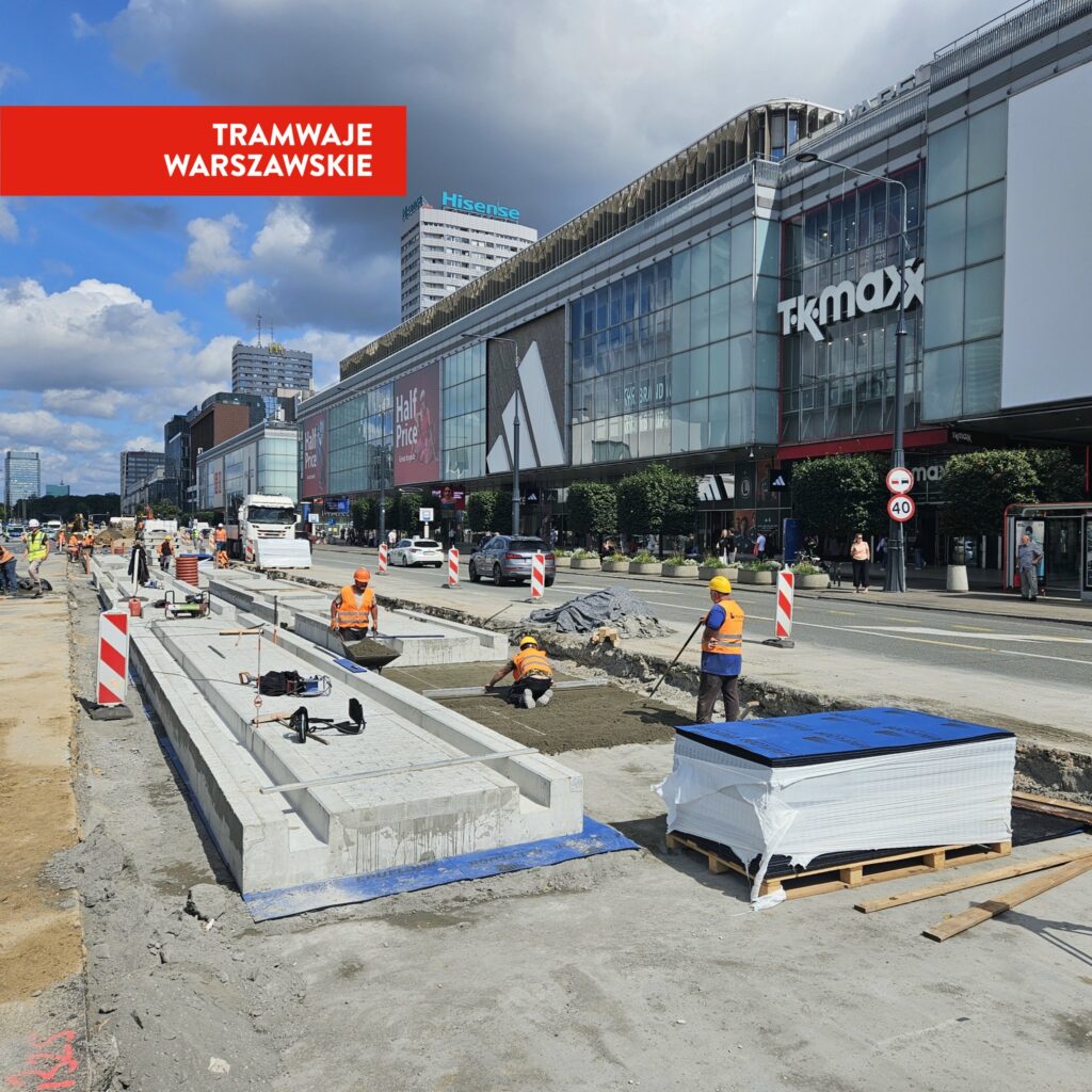 Grupa pracowników w pomarańczowych kamizelkach roboczych układa tory tramwajowe na miejskiej ulicy, obok nowoczesnych budynków.