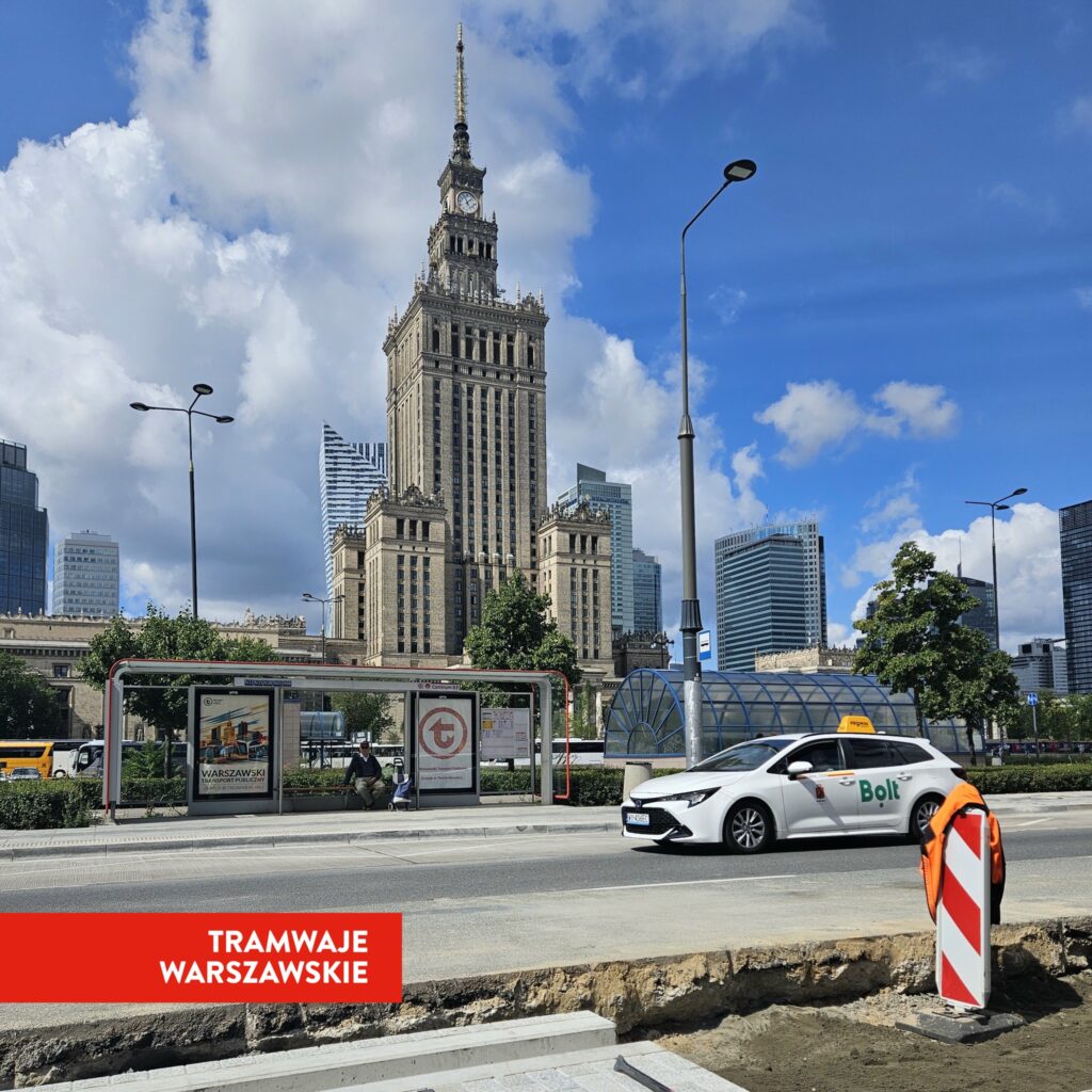 Biały samochód marki Bolt przejeżdża obok przystanku tramwajowego z nowoczesnym biurowcem w tle.